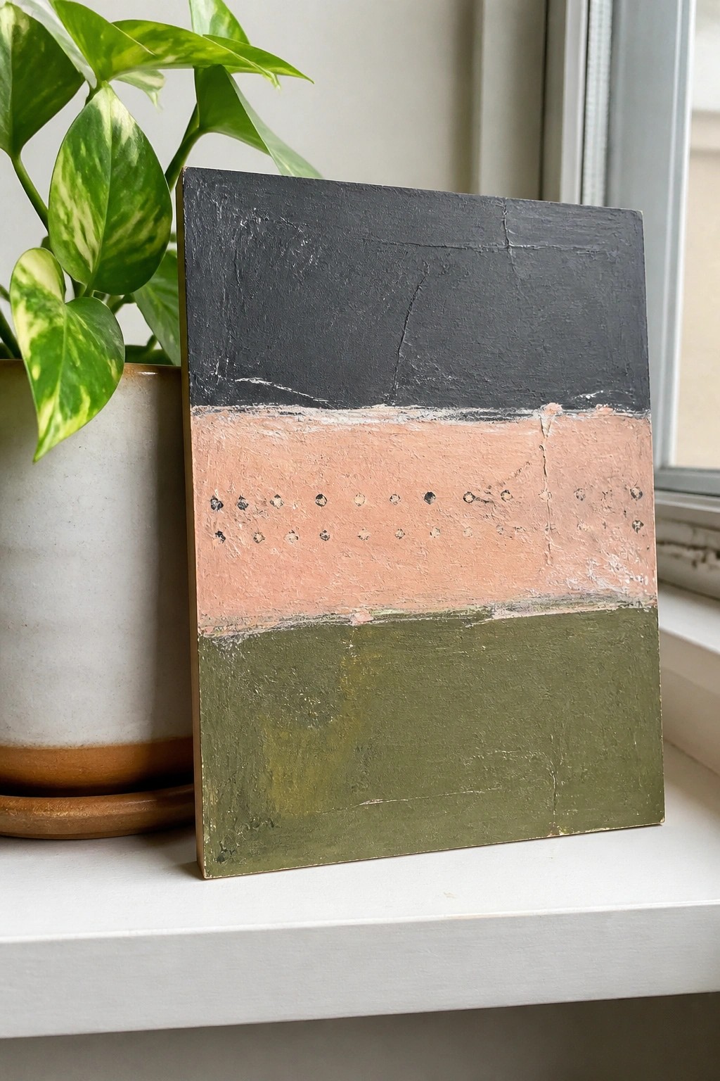 Abstract painting with black, peach, and green bands beside potted plant on windowsill.