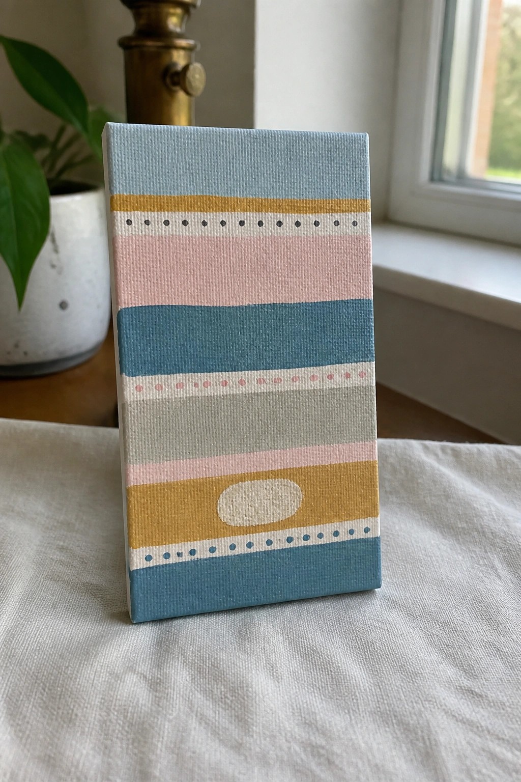 Small canvas with pastel stripes, dots, and oval on wooden surface near plant