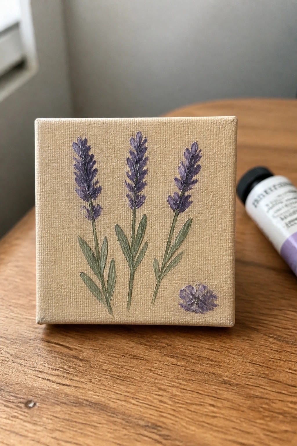 Small beige canvas with three purple lavender stems beside paint tube on wood table