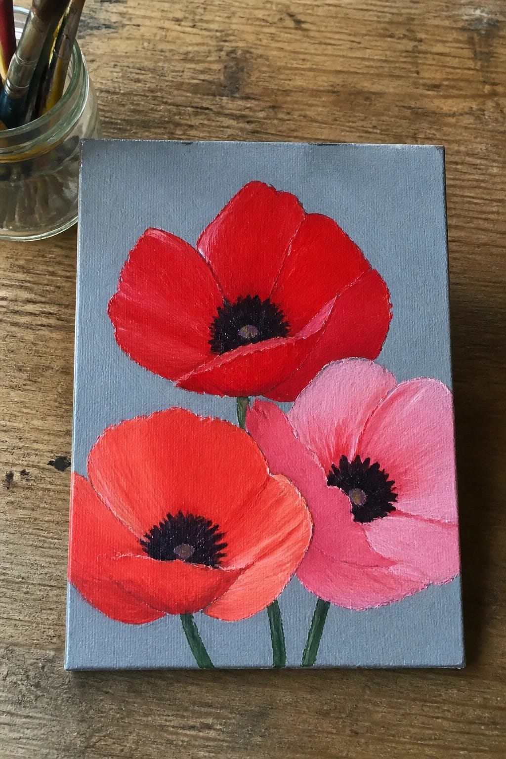 Red and pink poppies painted on small gray canvas beside paintbrushes on wood.