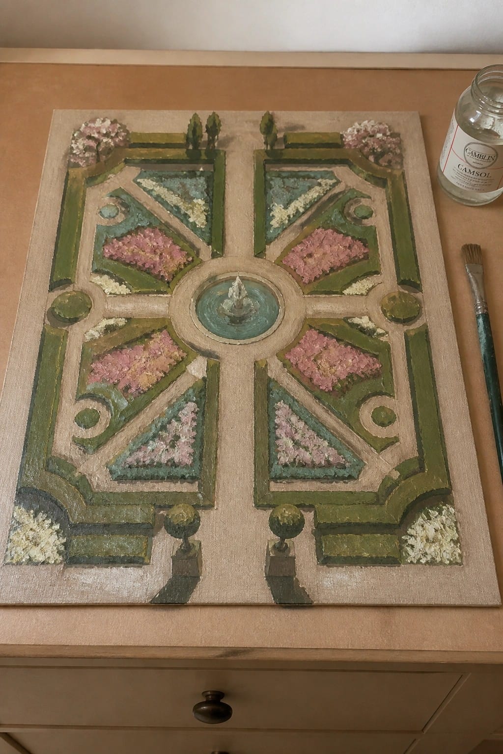 Oil painting of formal garden with central fountain, pink flower beds, and topiaries beside art supplies