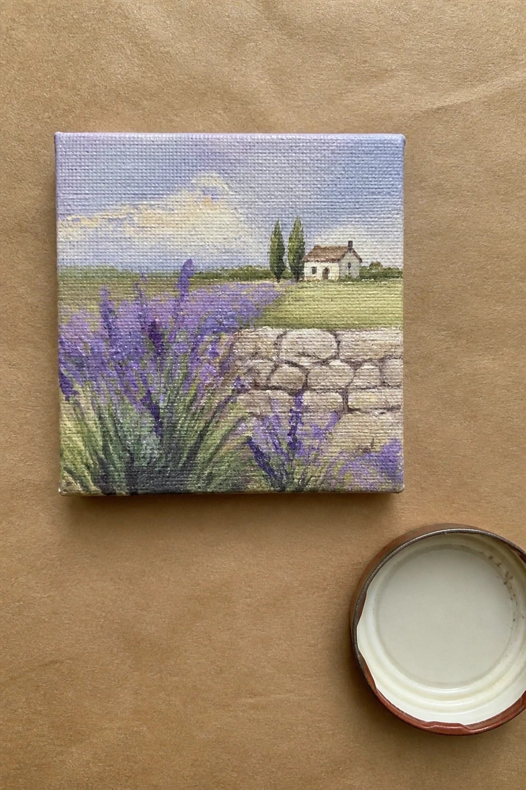 A small square oil painting of purple lavender flowers beside a stone wall with fields, a house, and trees under a light sky, placed on brown paper next to an open paint lid.