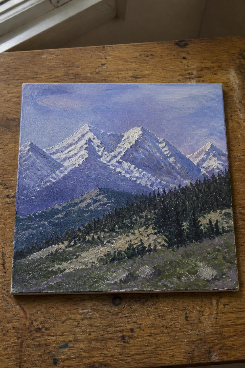 Square canvas painting of snow-capped mountains and green forest on wooden table