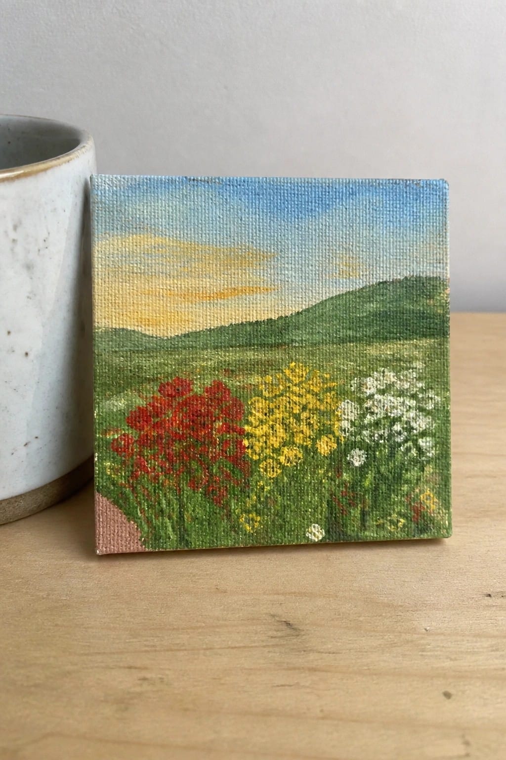 Small square landscape painting of red, yellow, and white wildflowers beside a mug.
