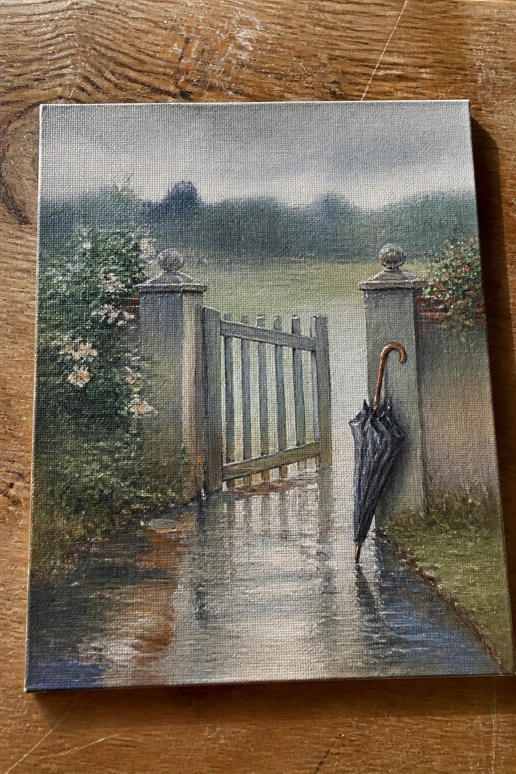 Realistic painting of rainy path with open gate, leaning black umbrella, and puddles.