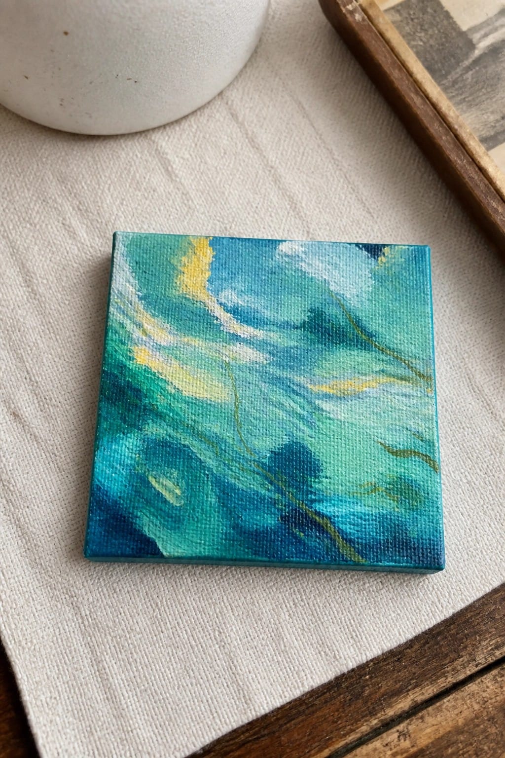 Small square canvas with swirling blue, green, and yellow abstract painting on white fabric