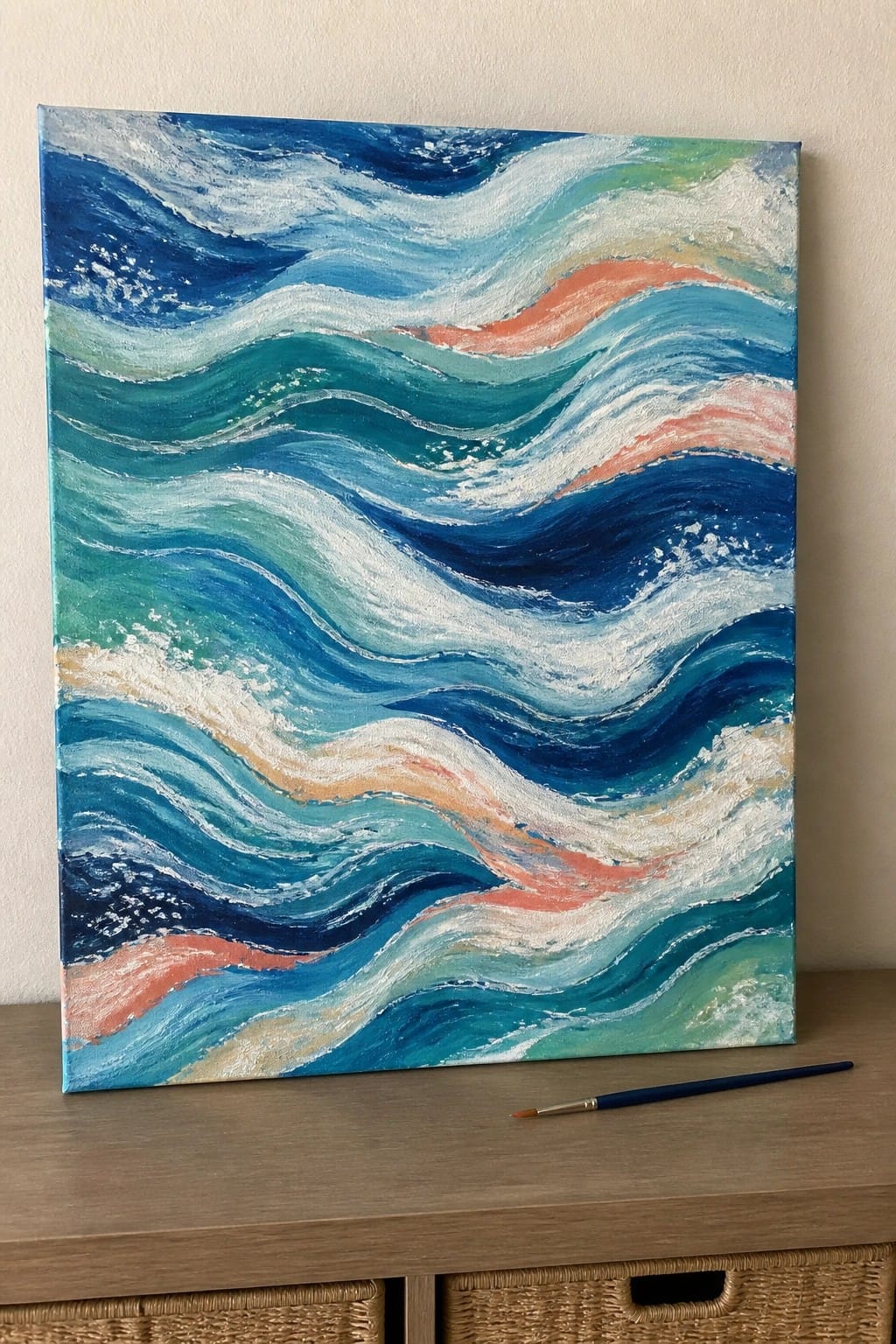 Abstract wave painting in blues, teals, and coral on canvas beside paintbrush on wood
