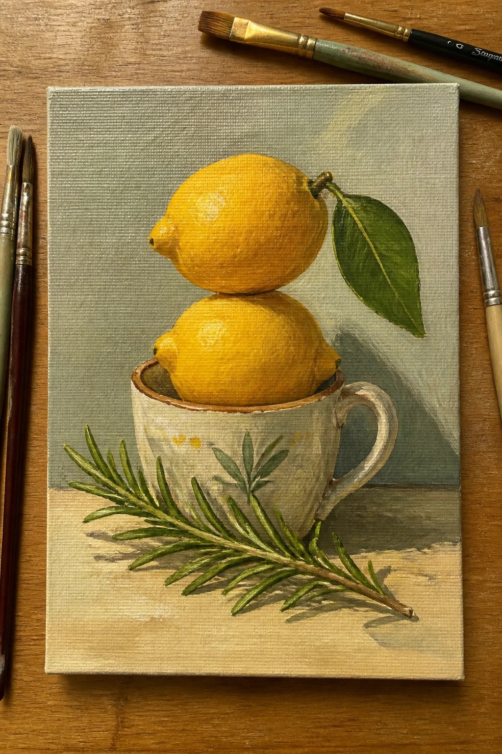 Still life painting of two lemons in a white cup with rosemary and brushes