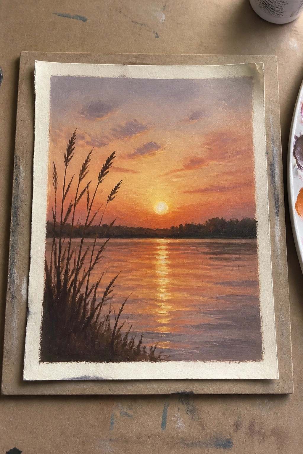 Sunset painting with orange sky, glowing sun reflection on water, silhouetted reeds.
