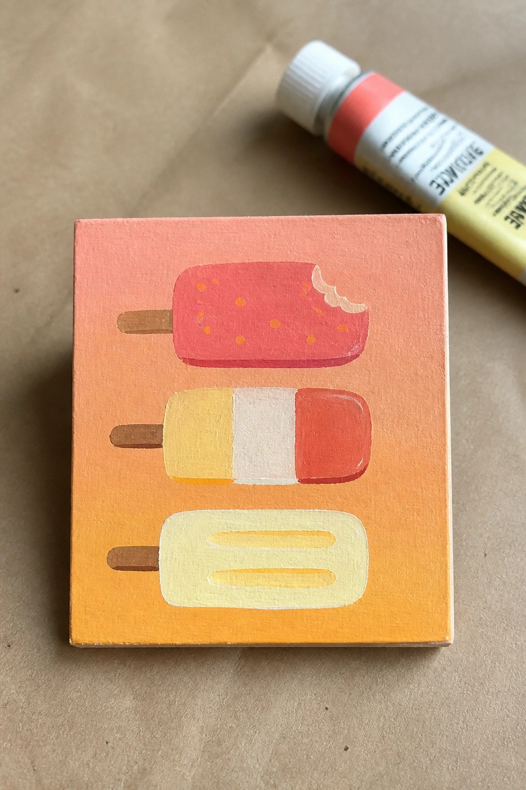 Small canvas with three colorful popsicles on orange-pink gradient beside paint tube