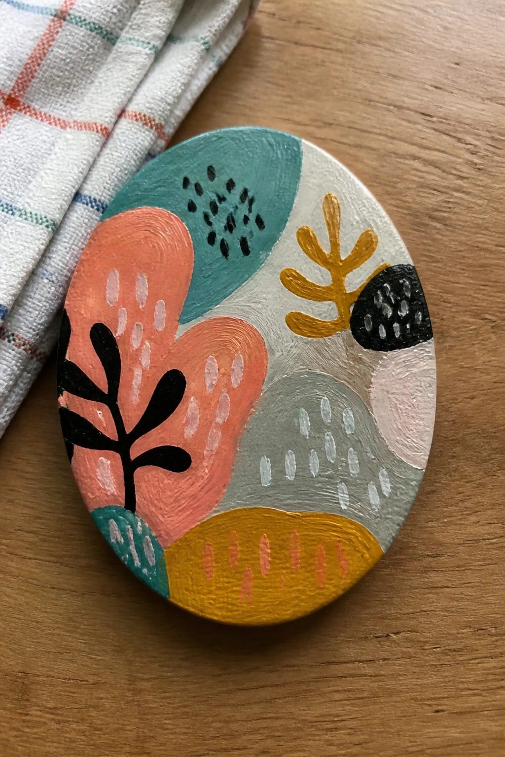 Colorful abstract painted oval stone on wooden table beside checkered cloth
