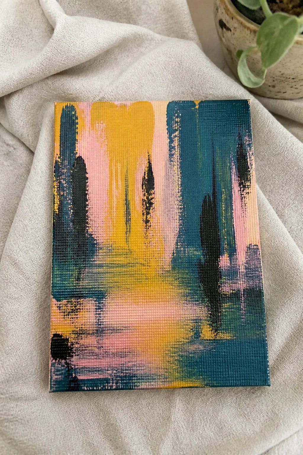 Abstract painting with vertical teal, yellow, pink brushstrokes on white fabric beside potted plant.