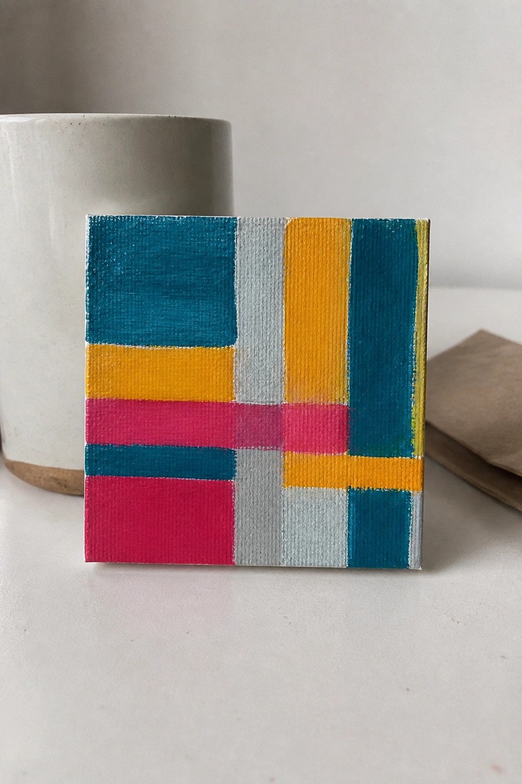 Small colorful geometric abstract painting beside white ceramic mug on table