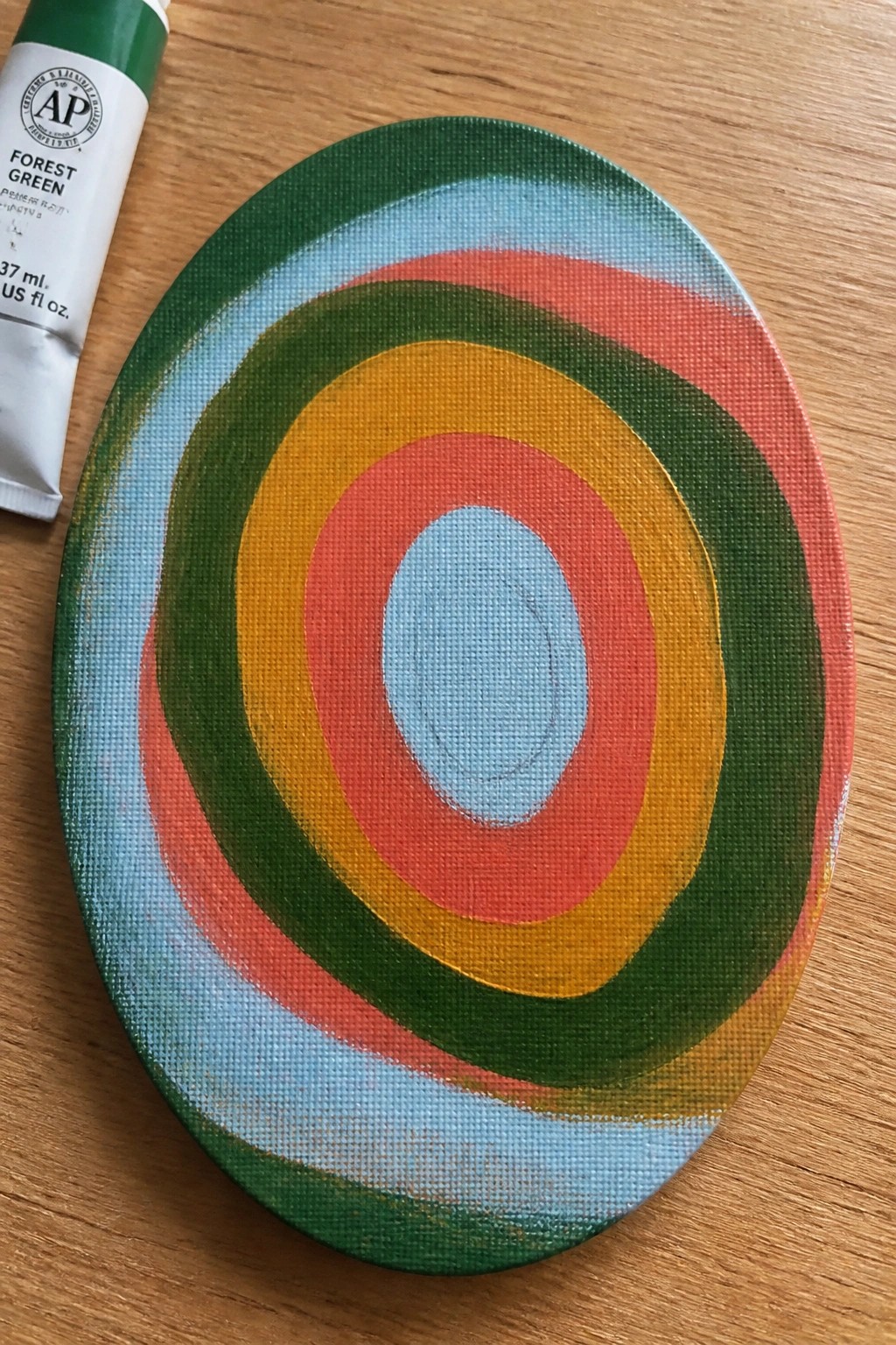 Oval canvas with colorful concentric rings beside forest green paint tube on wood.