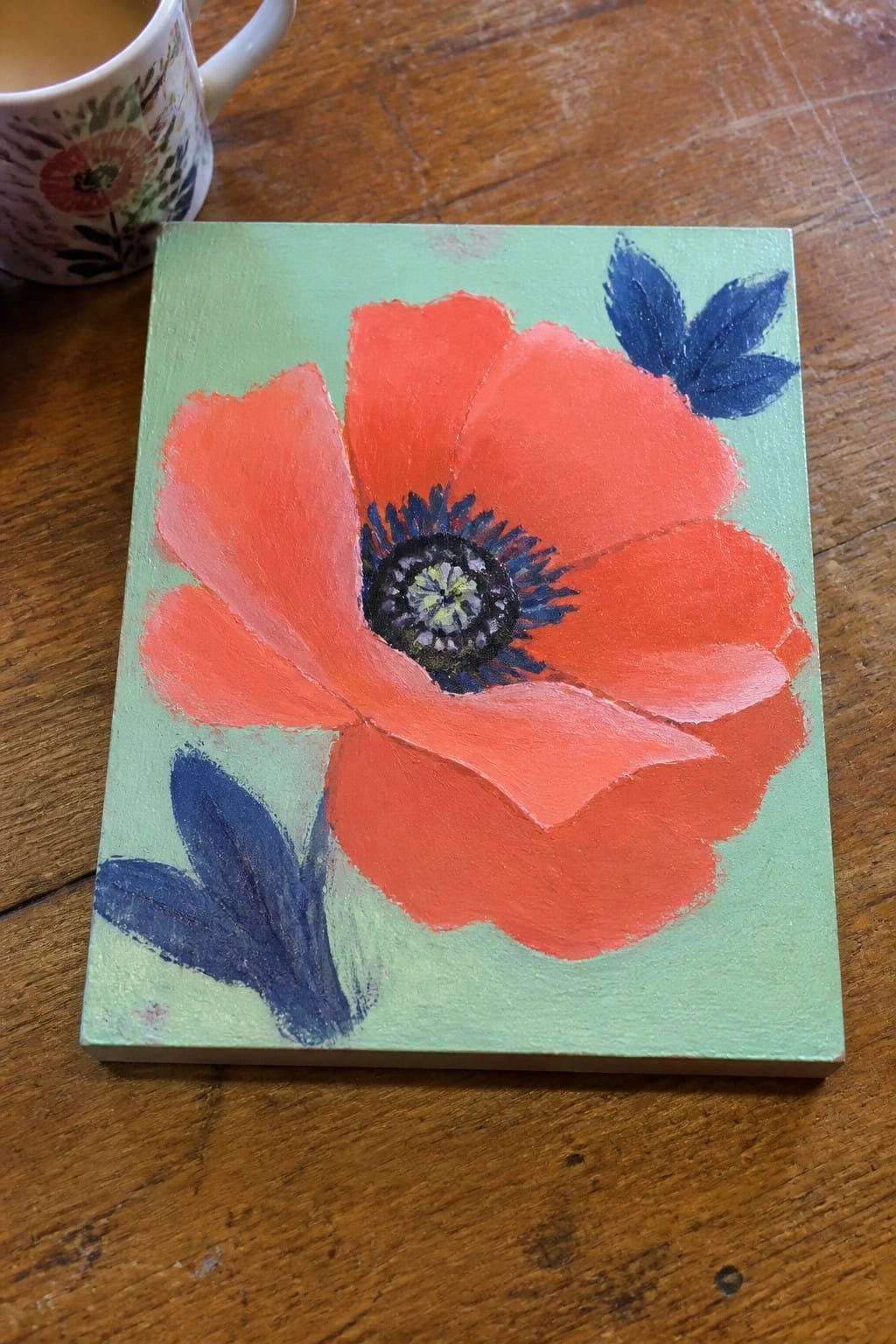 Red poppy with blue leaves painted on green canvas beside floral mug on wood.