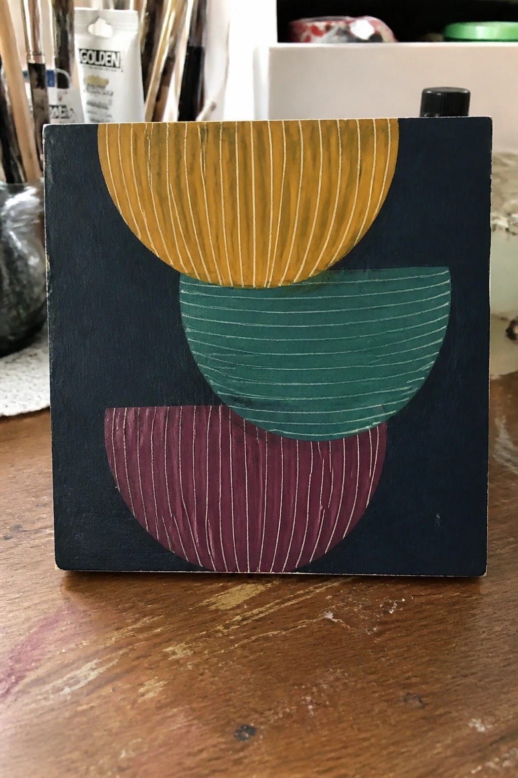 An abstract oil painting with three overlapping half-circles in yellow, teal, and maroon, each with white vertical stripes, on a dark blue background.