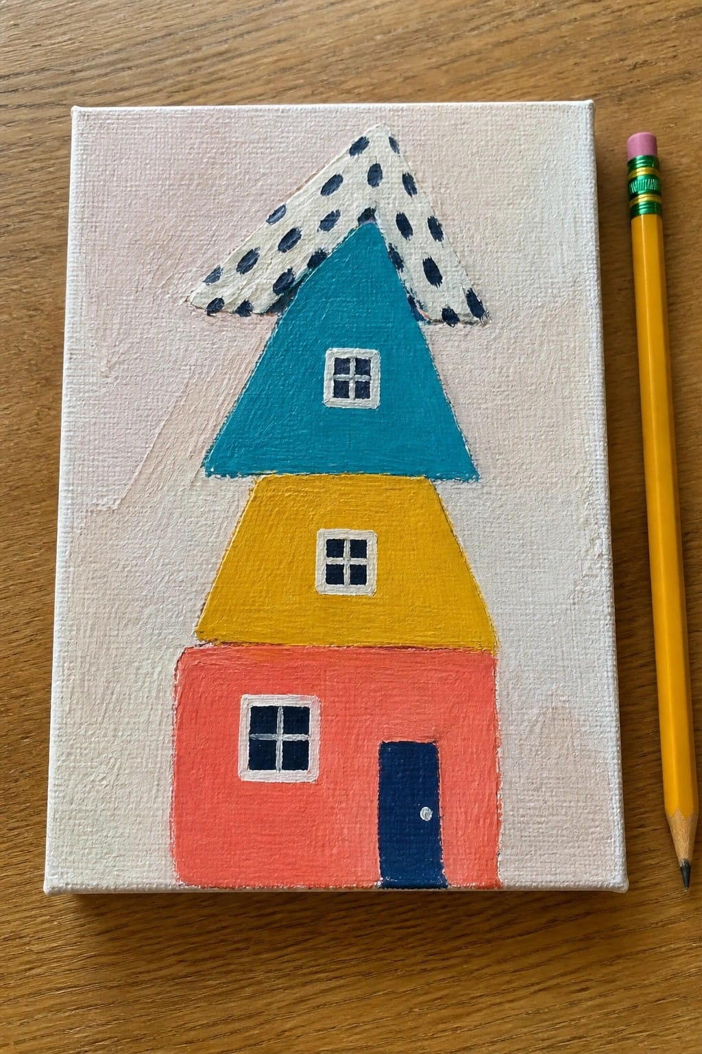 Colorful stacked house painting on canvas with red yellow teal and dotted roof beside pencil