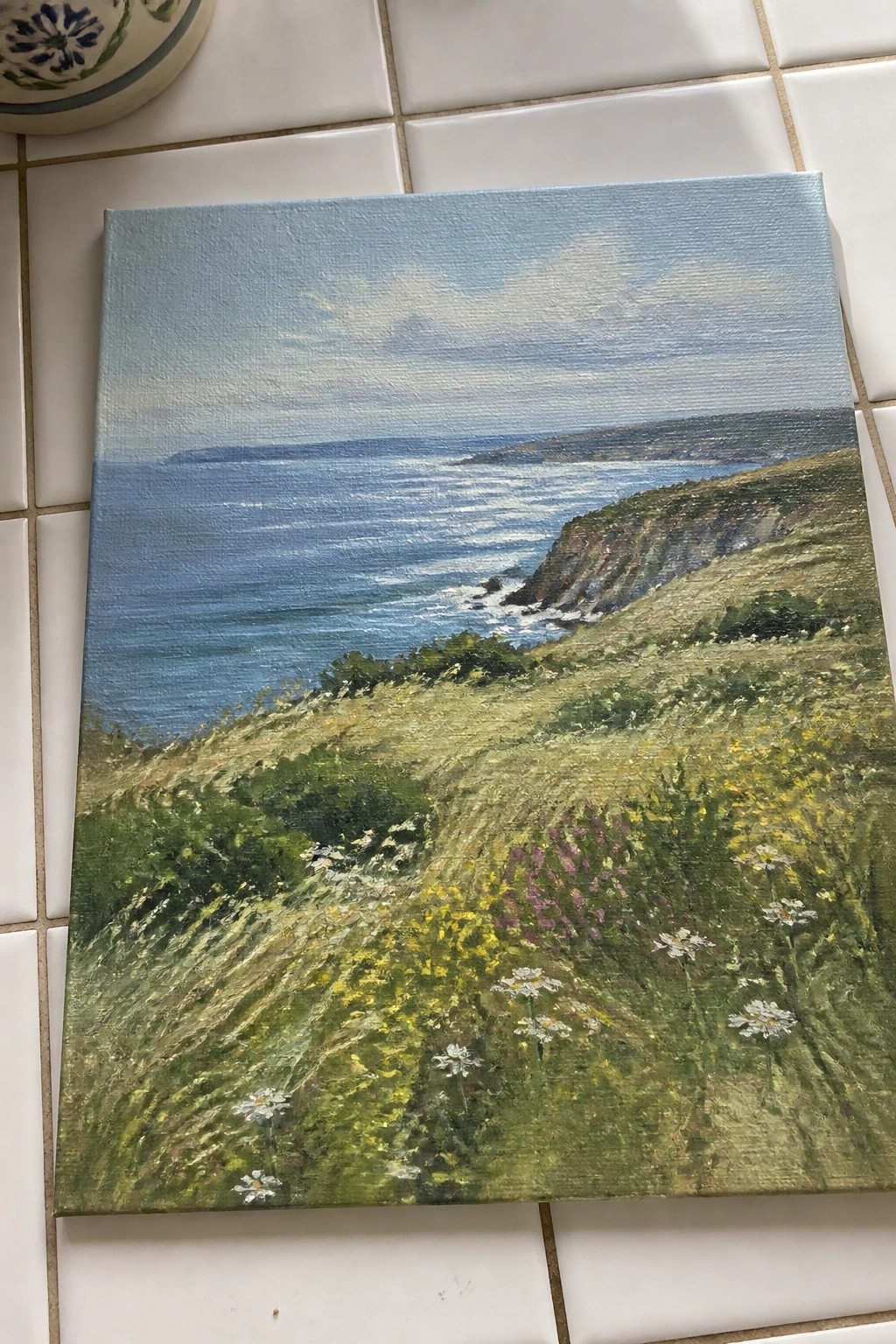 Oil painting of ocean waves, rocky cliffs, and wildflower meadow under blue sky