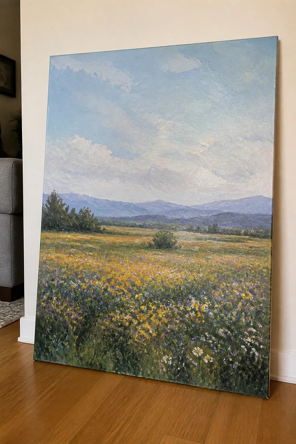 An oil painting depicts a vibrant meadow filled with yellow, purple, and white wildflowers leading to distant mountains under a blue sky with clouds.