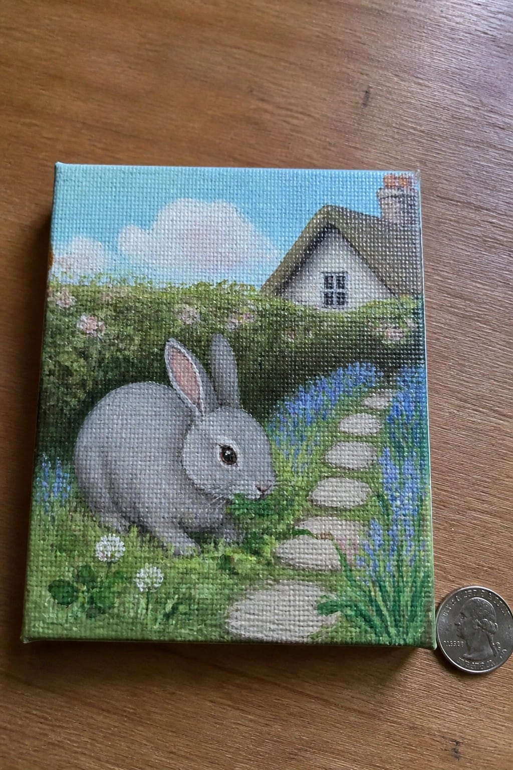Miniature canvas painting of gray rabbit grazing in garden by cottage, beside quarter.