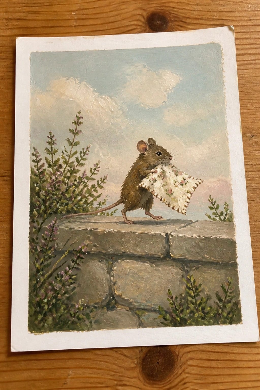 Brown mouse on stone wall holds floral cloth amid wildflowers under cloudy sky
