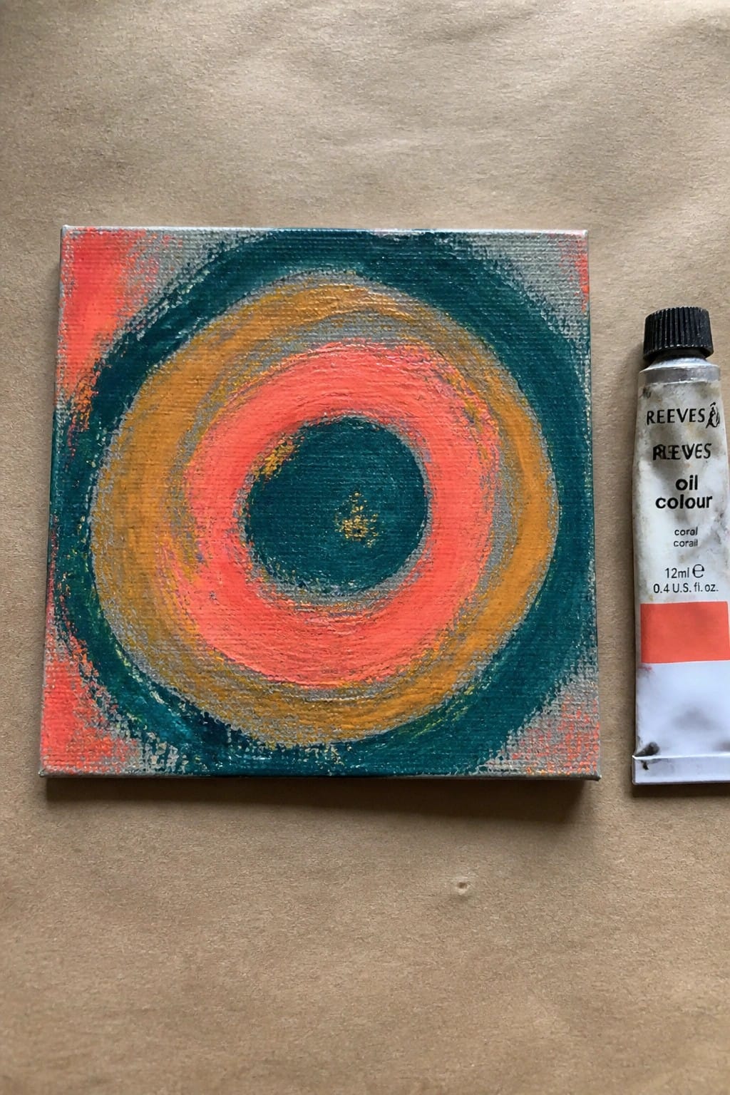 A small square oil painting of concentric circles in teal, orange, coral, and yellow on a canvas next to a tube of coral oil paint.