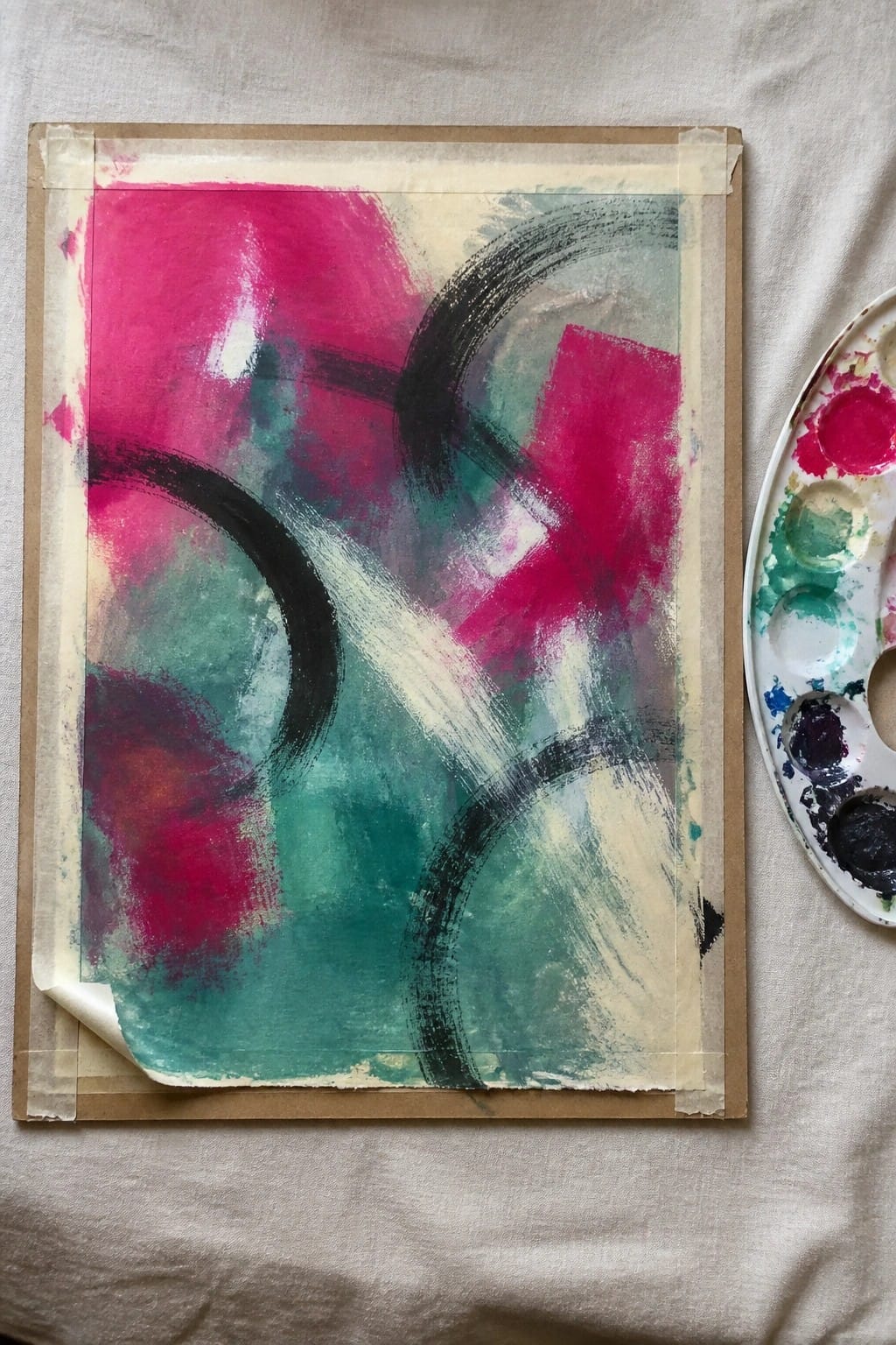 Abstract painting with bold pink, teal, and black strokes beside paint palette on fabric