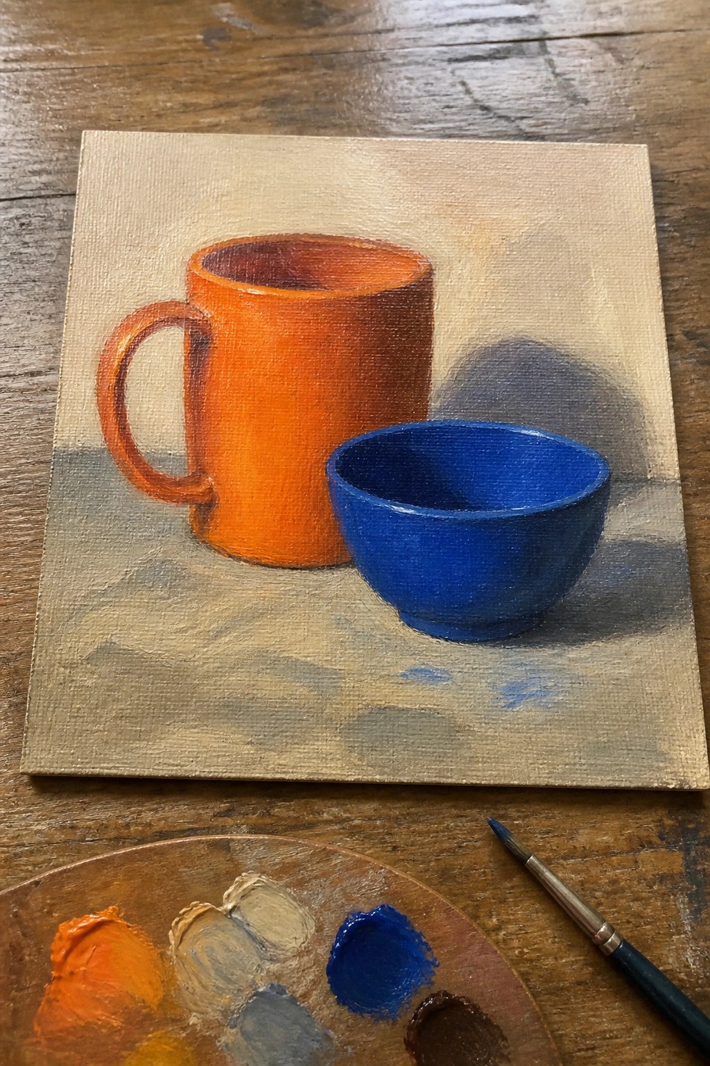 An oil painting of an orange mug and blue bowl on a neutral surface with a paint palette and brush in the foreground.