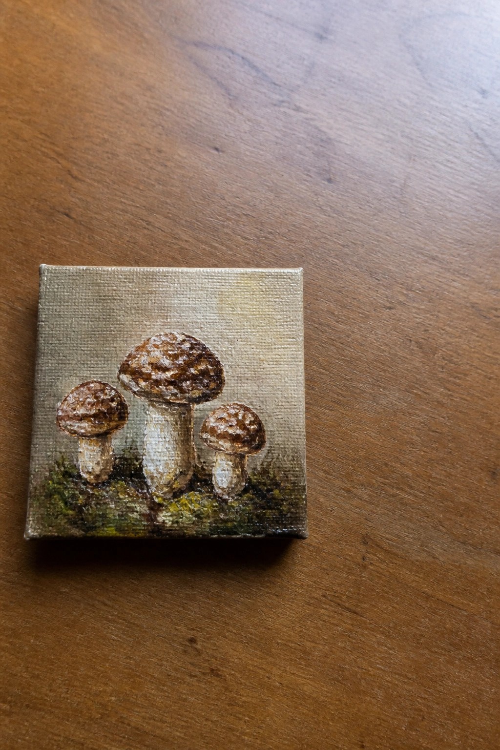 Small square canvas painting of three brown mushrooms on wooden table.