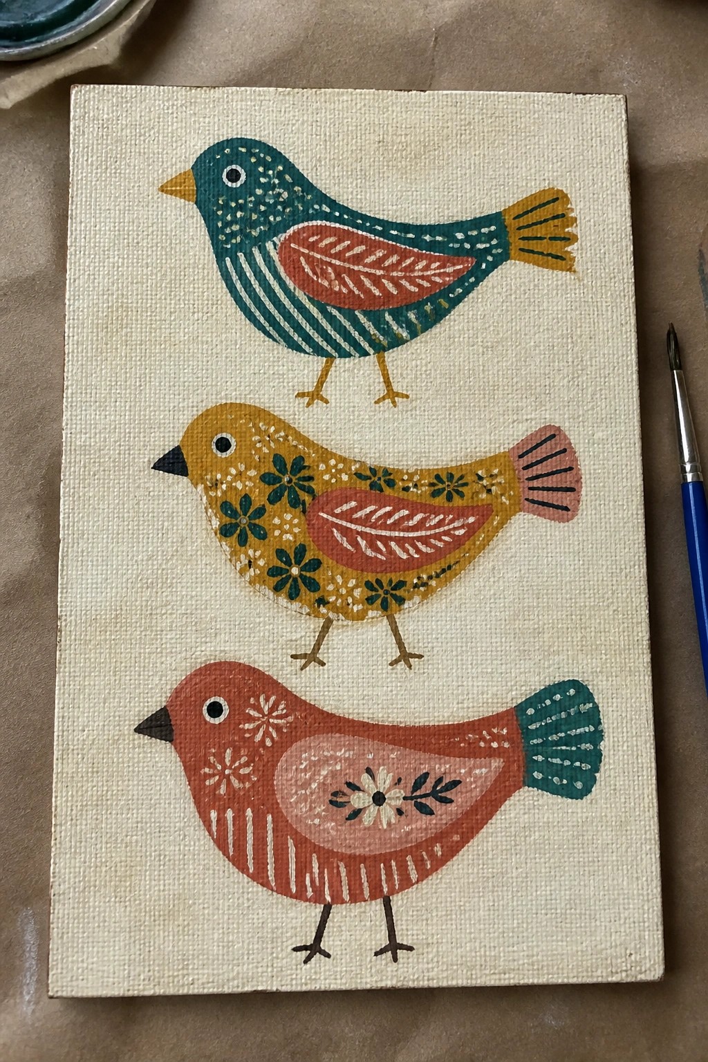 Three folk-art birds in teal, yellow, and red with patterns on canvas beside brush.