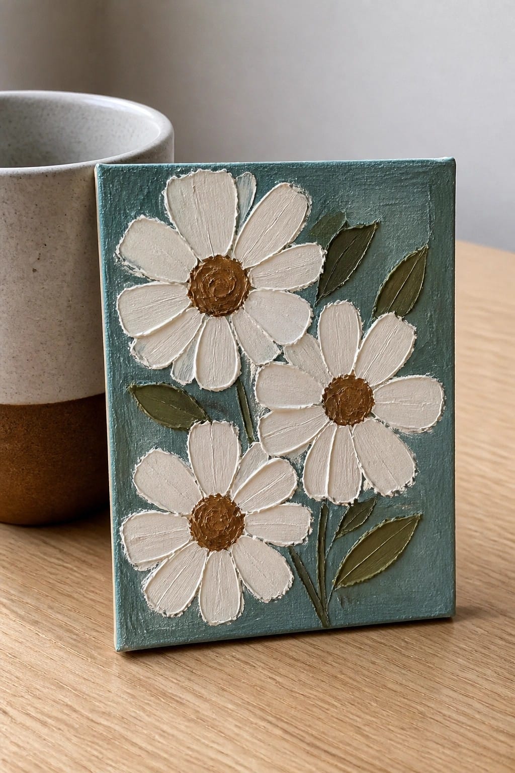 Textured painting of three white daisies with brown centers on teal background beside ceramic mug.