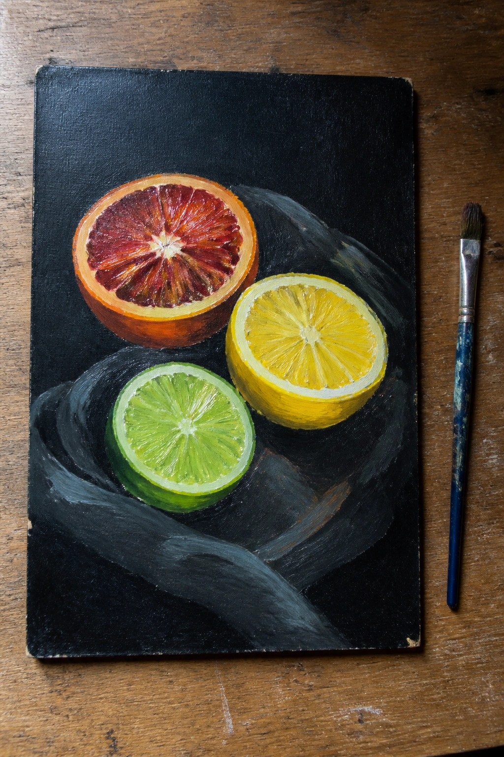 Black canvas still life with blood orange, lemon, lime halves and paintbrush on wood
