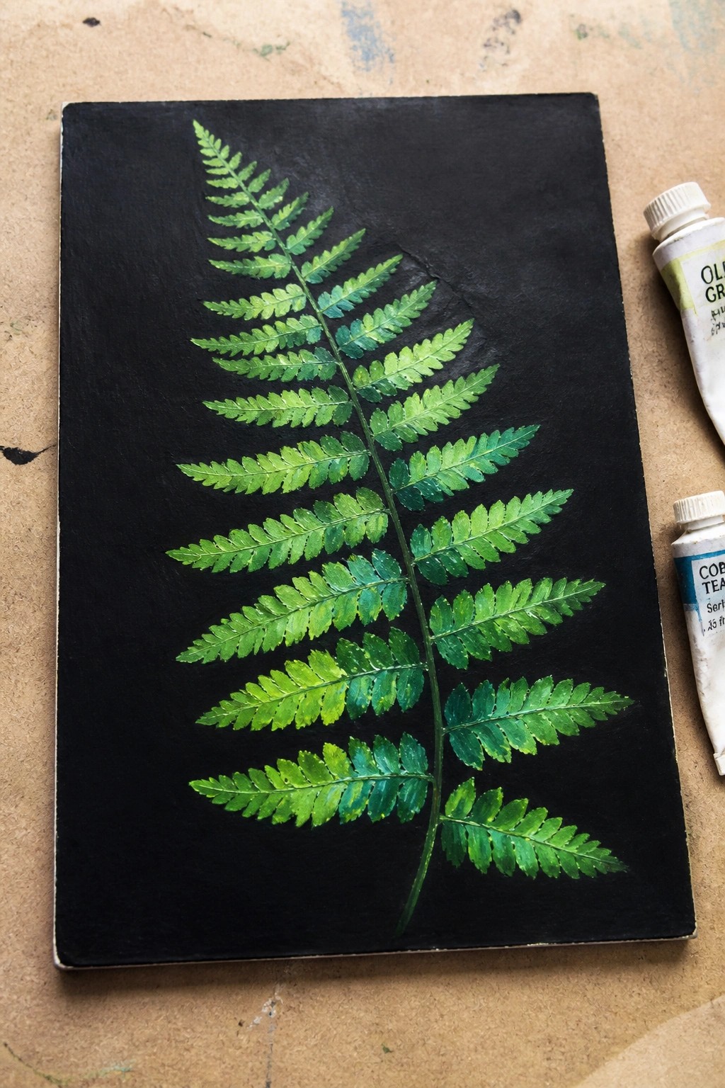 Painted green fern frond on black paper beside two paint tubes.