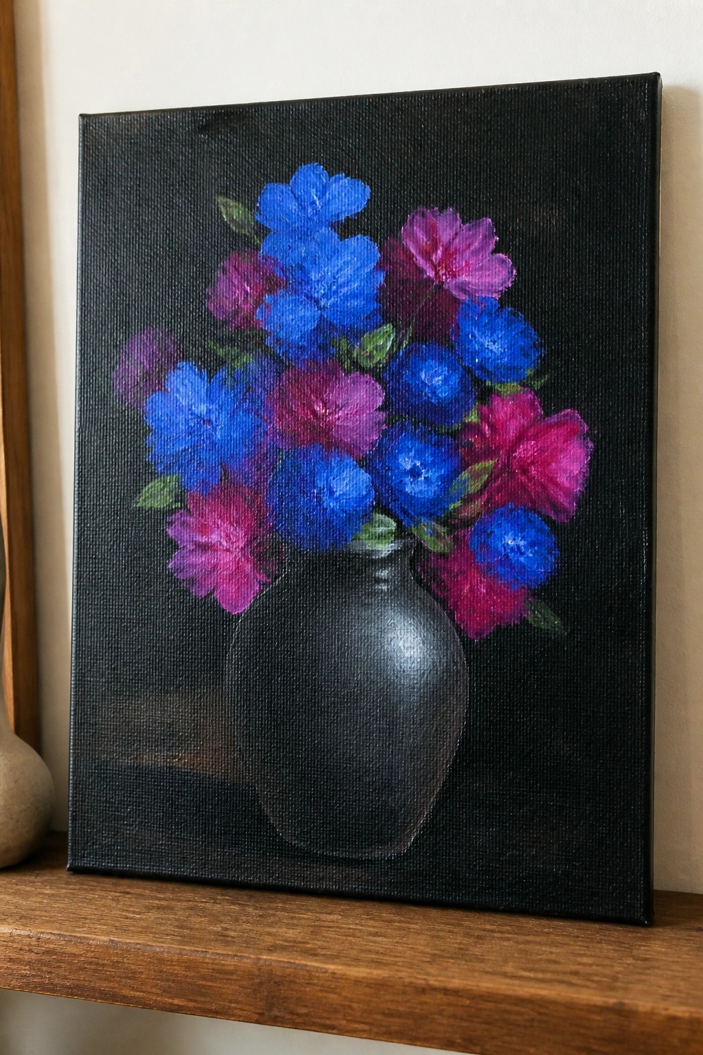 Painting of blue and magenta flowers in a black vase on wooden shelf.