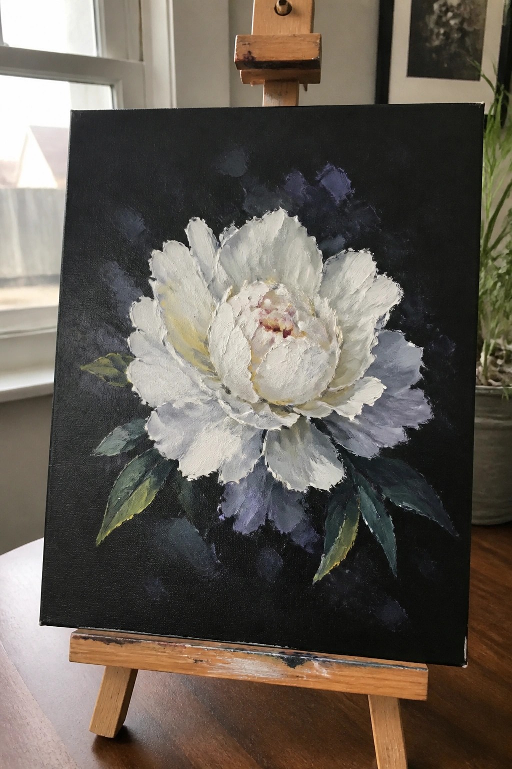 White peony oil painting with green leaves on black canvas atop wooden easel