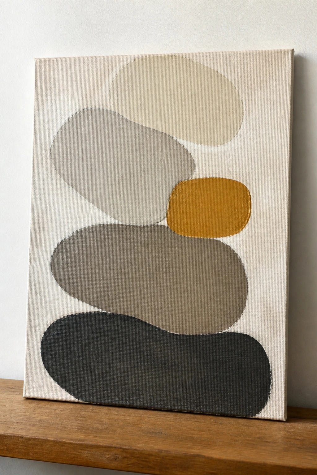 Abstract canvas with overlapping ovals in neutral tones and mustard yellow on wood ledge.