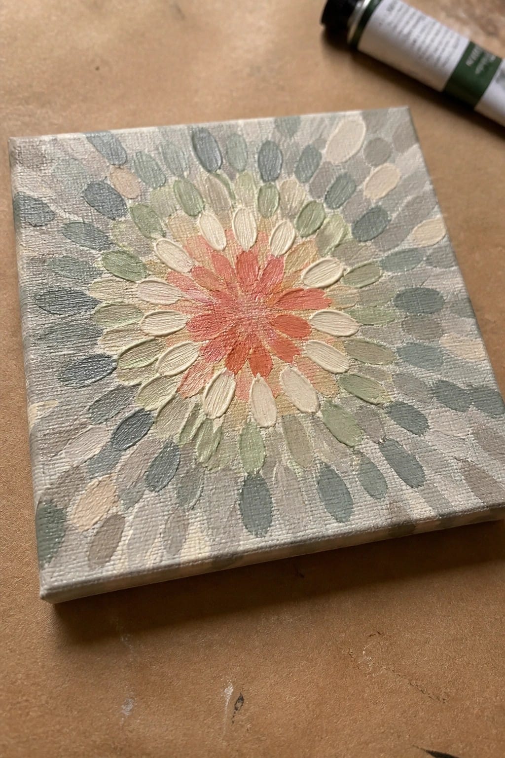 Thickly painted coral flower mandala on square canvas beside green paint tube.