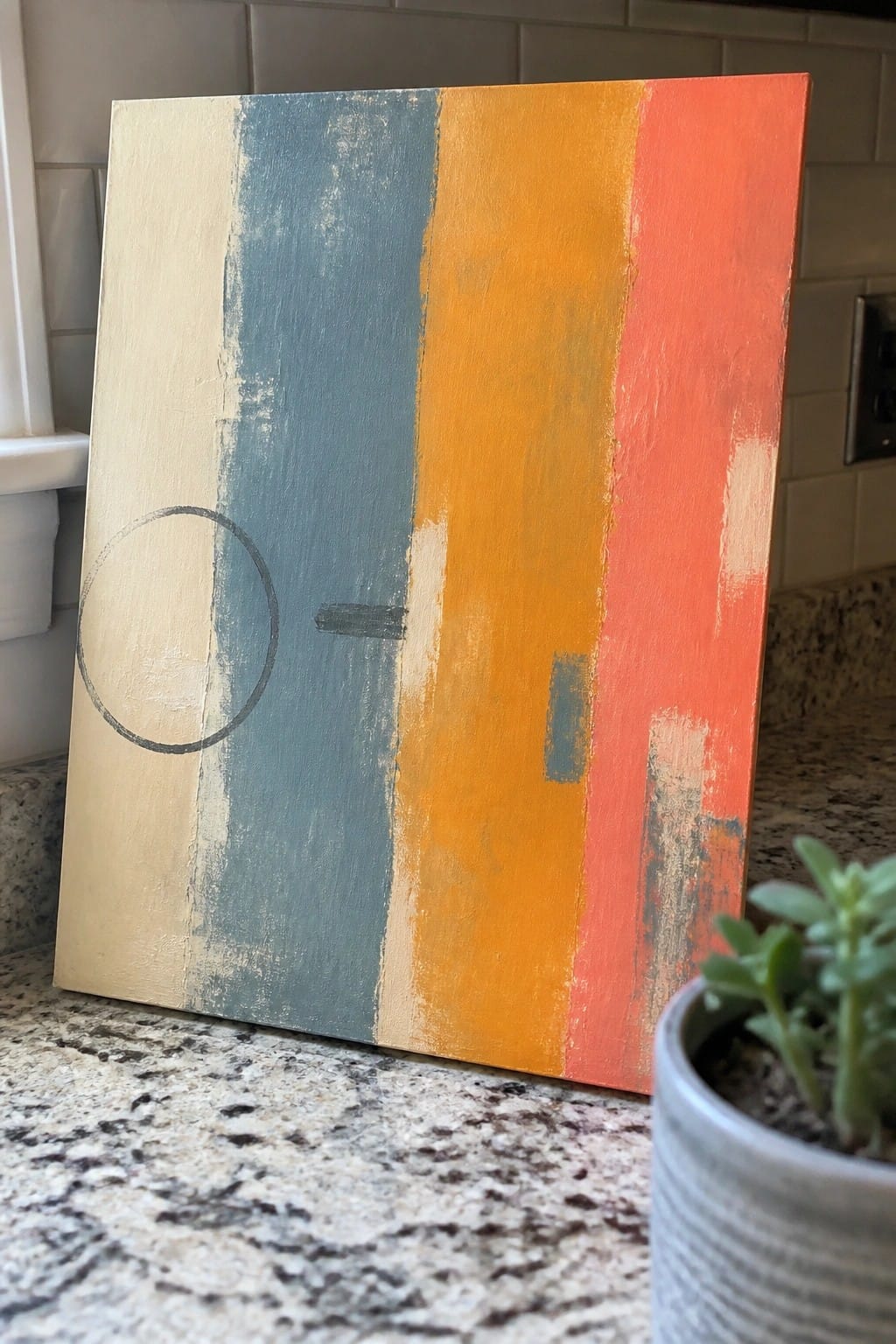 Abstract canvas with beige, blue, orange, and red stripes plus circle and dash on countertop beside plant