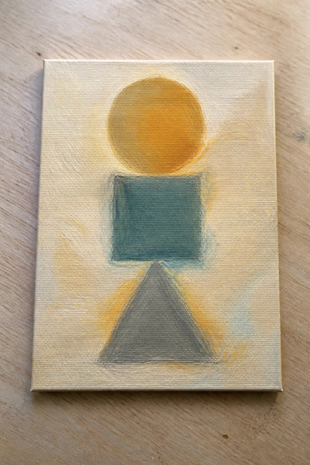 Abstract painting with yellow circle above teal square and gray triangle on pale canvas.