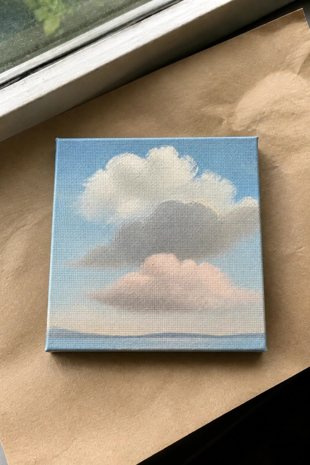 Small square canvas painting of fluffy white and gray clouds in blue sky on brown paper.