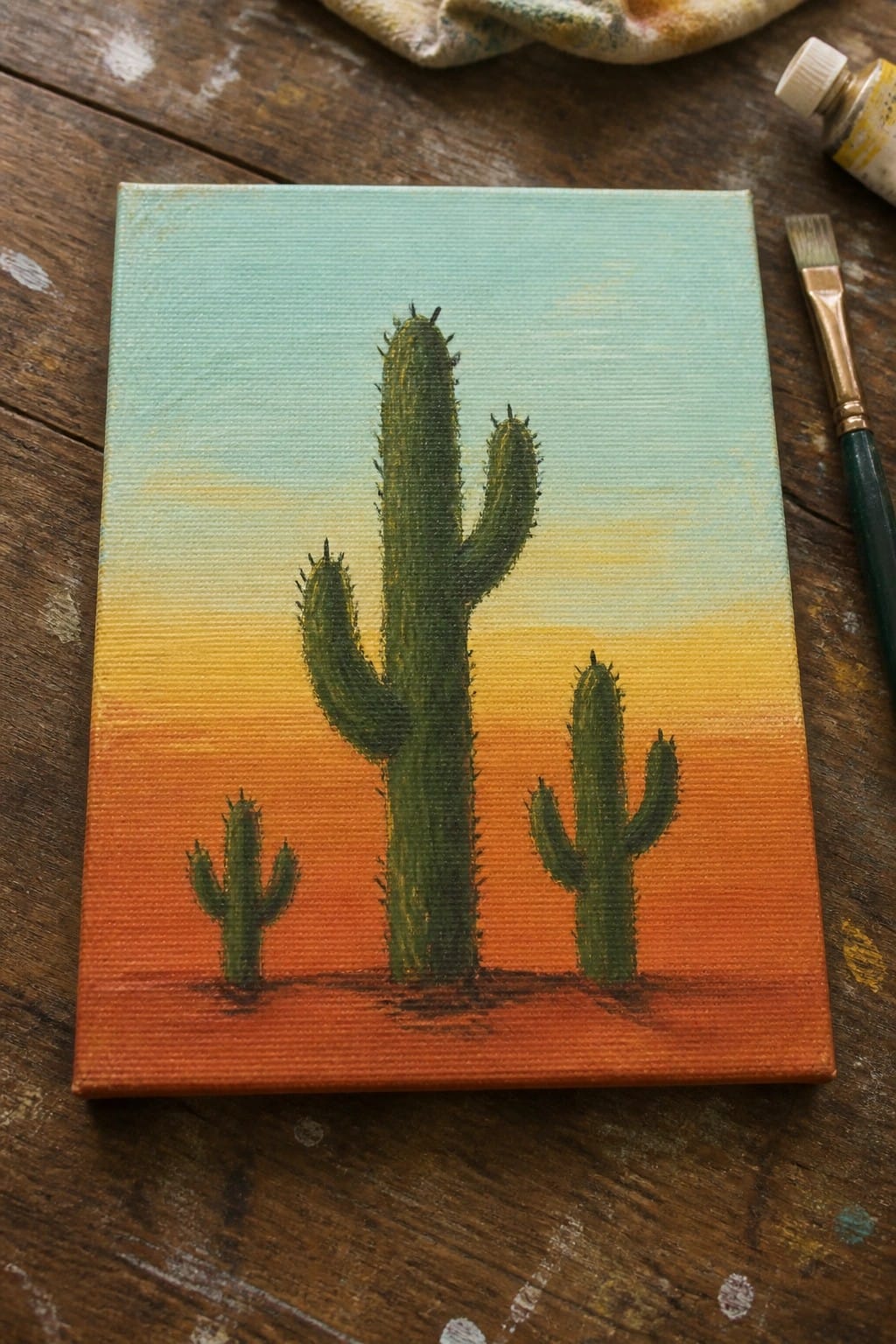 A small canvas shows green cacti of different sizes painted against a gradient sunset sky from blue to orange over reddish ground.