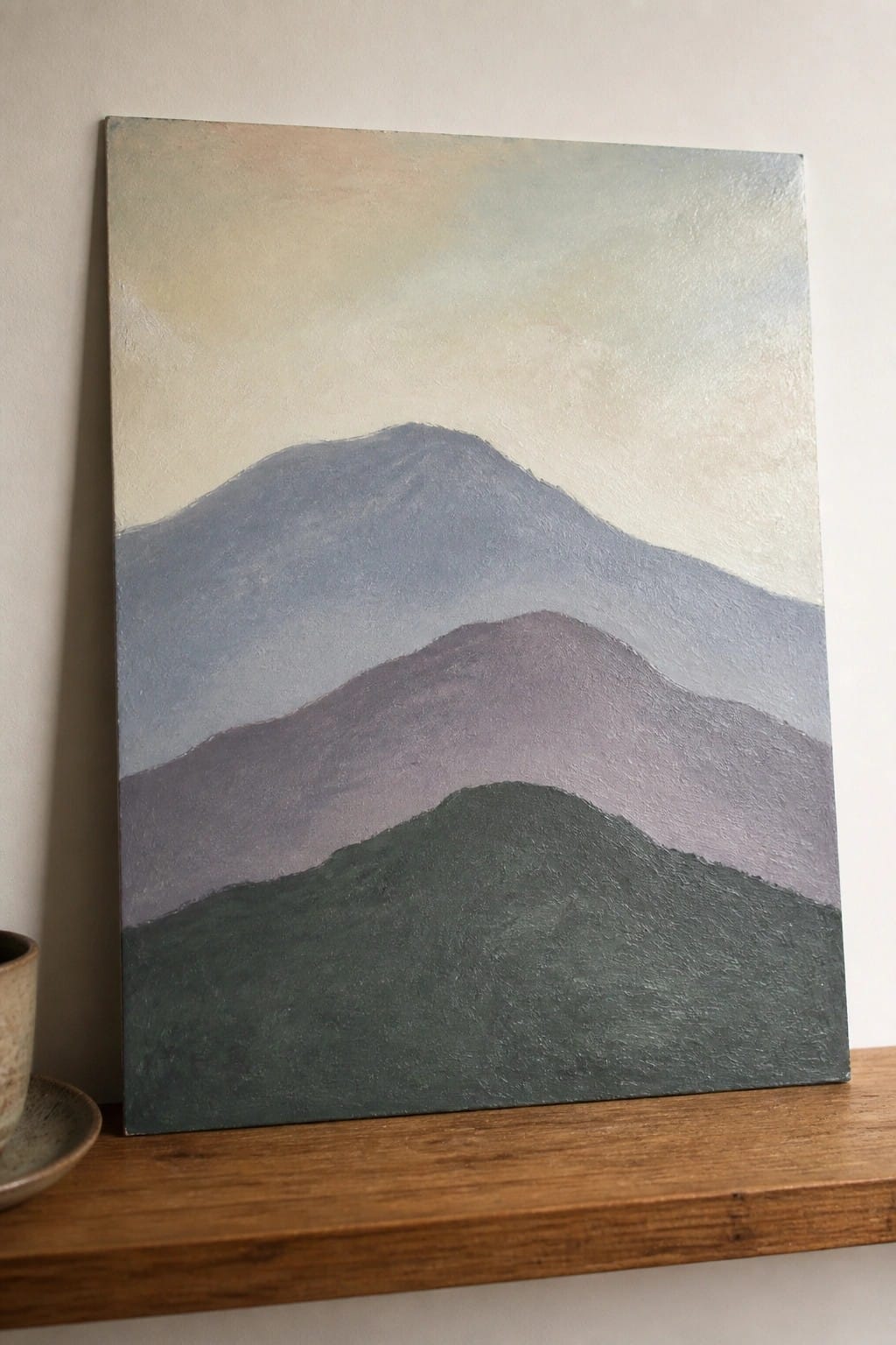 Layered abstract mountain painting in green, purple, blue on wooden shelf with cup