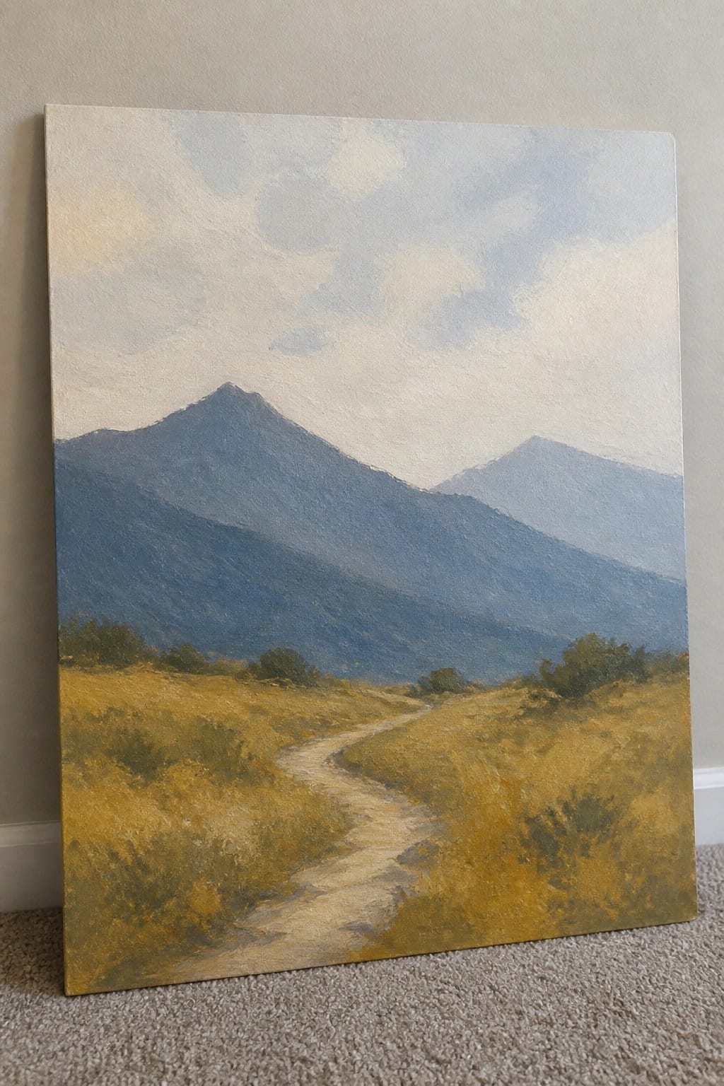 Winding dirt path through golden fields toward hazy blue mountains under cloudy sky.
