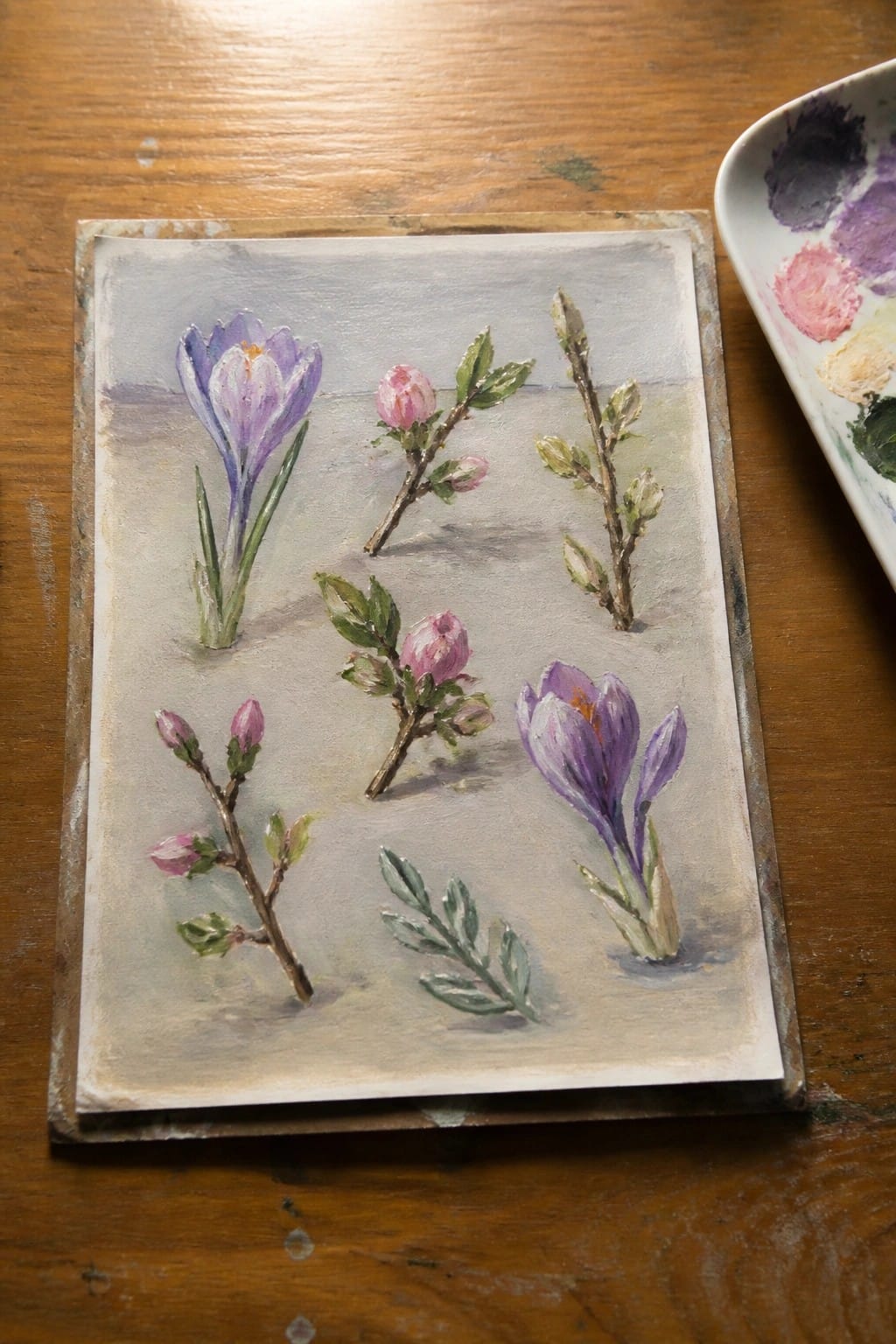 Watercolor painting of purple crocuses and pink buds beside paint palette on wood