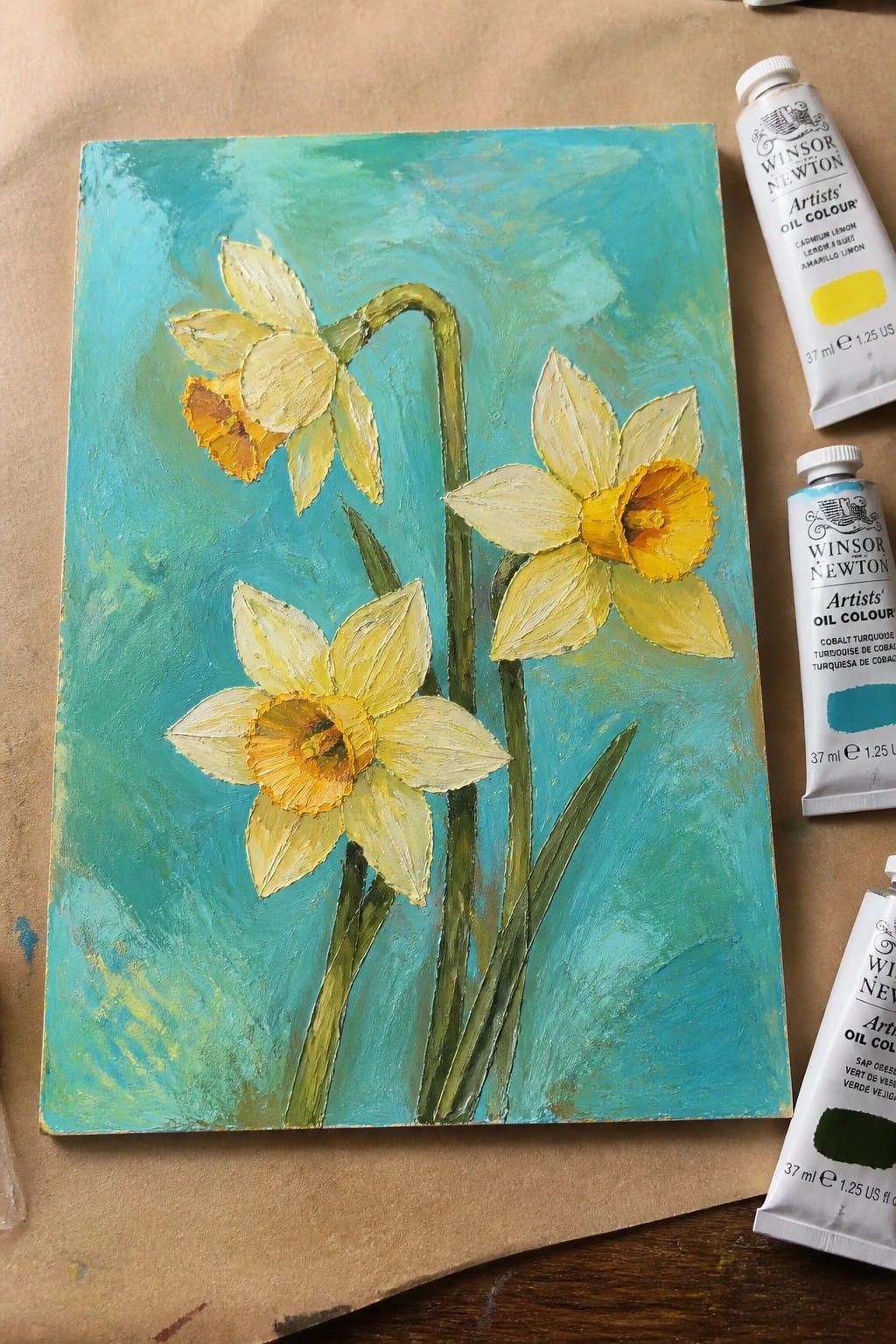 An oil painting of three yellow daffodils with orange centers against a teal background, shown next to tubes of lemon yellow, cobalt turquoise, and sap green paint.
