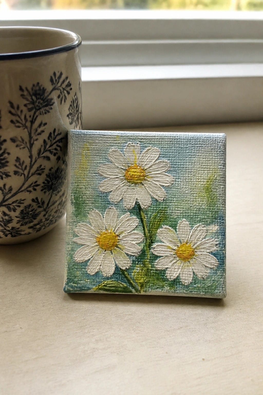 A small square oil painting of three white daisies with yellow centers and green stems on a blue-green background next to a floral mug.