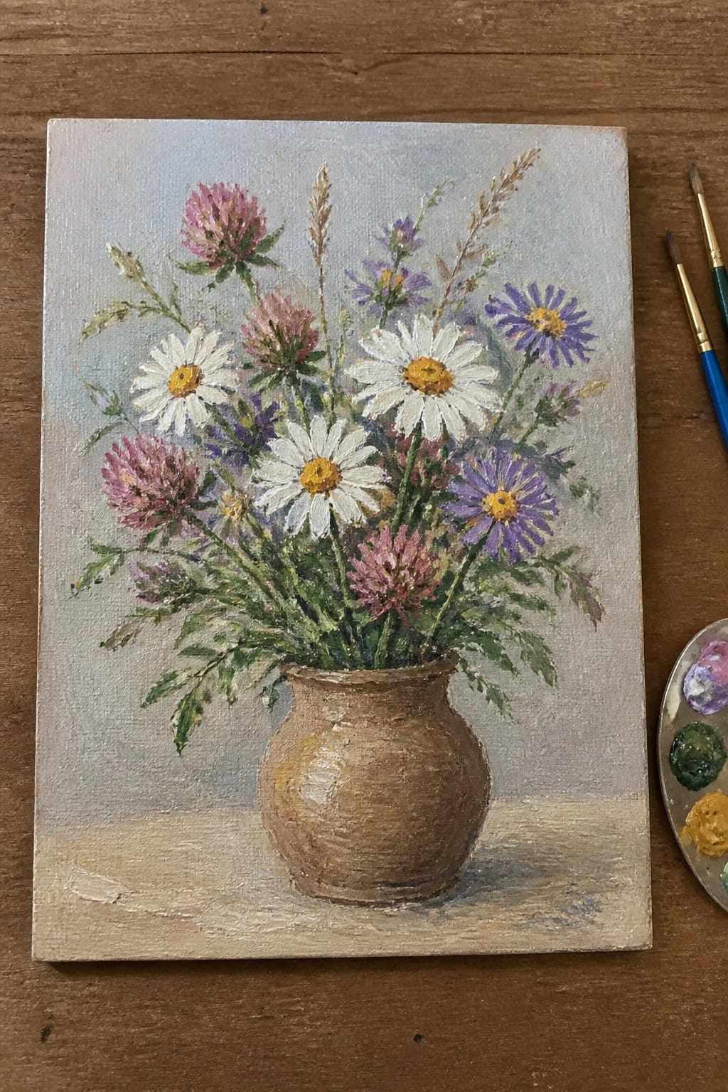 An oil painting of a brown clay vase holding white daisies, purple asters, and pink clover on a wooden surface next to paintbrushes and a palette.