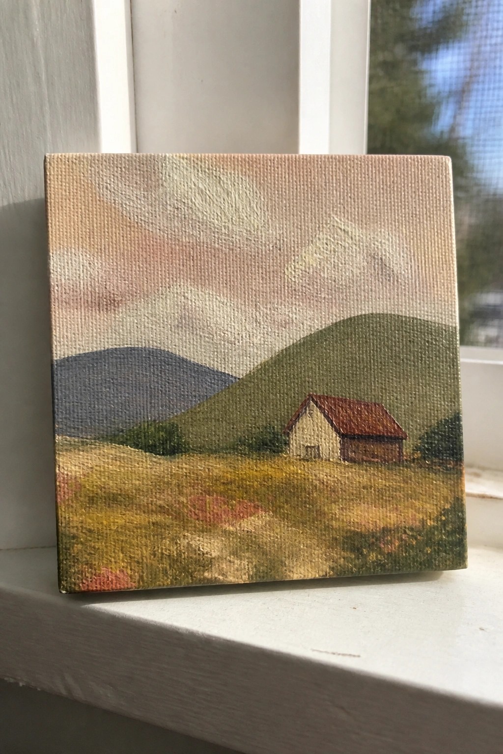 Small landscape painting of house with red roof amid green hills on white windowsill.