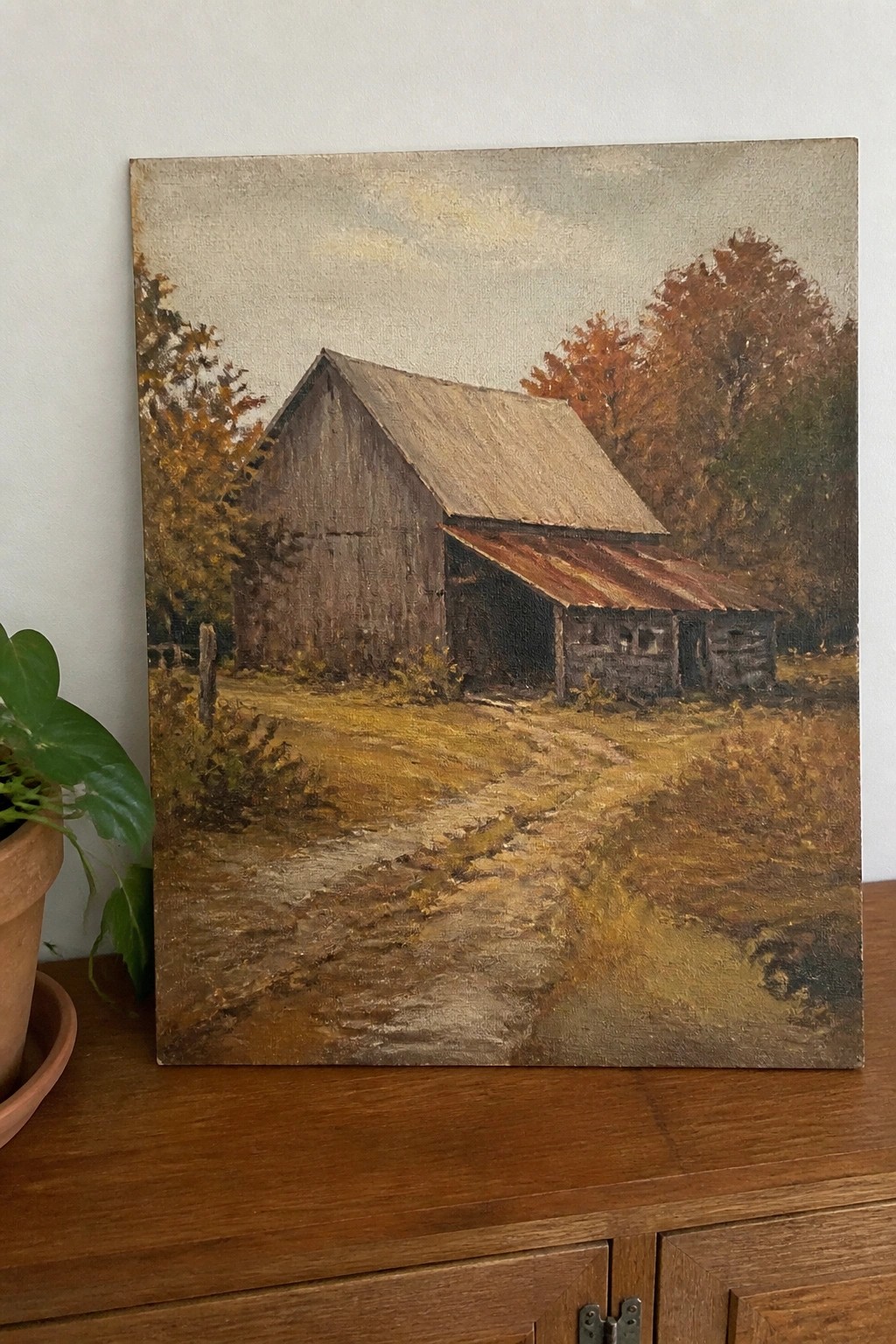 Weathered barn painting with rusty roof and autumn trees on wooden cabinet beside plant