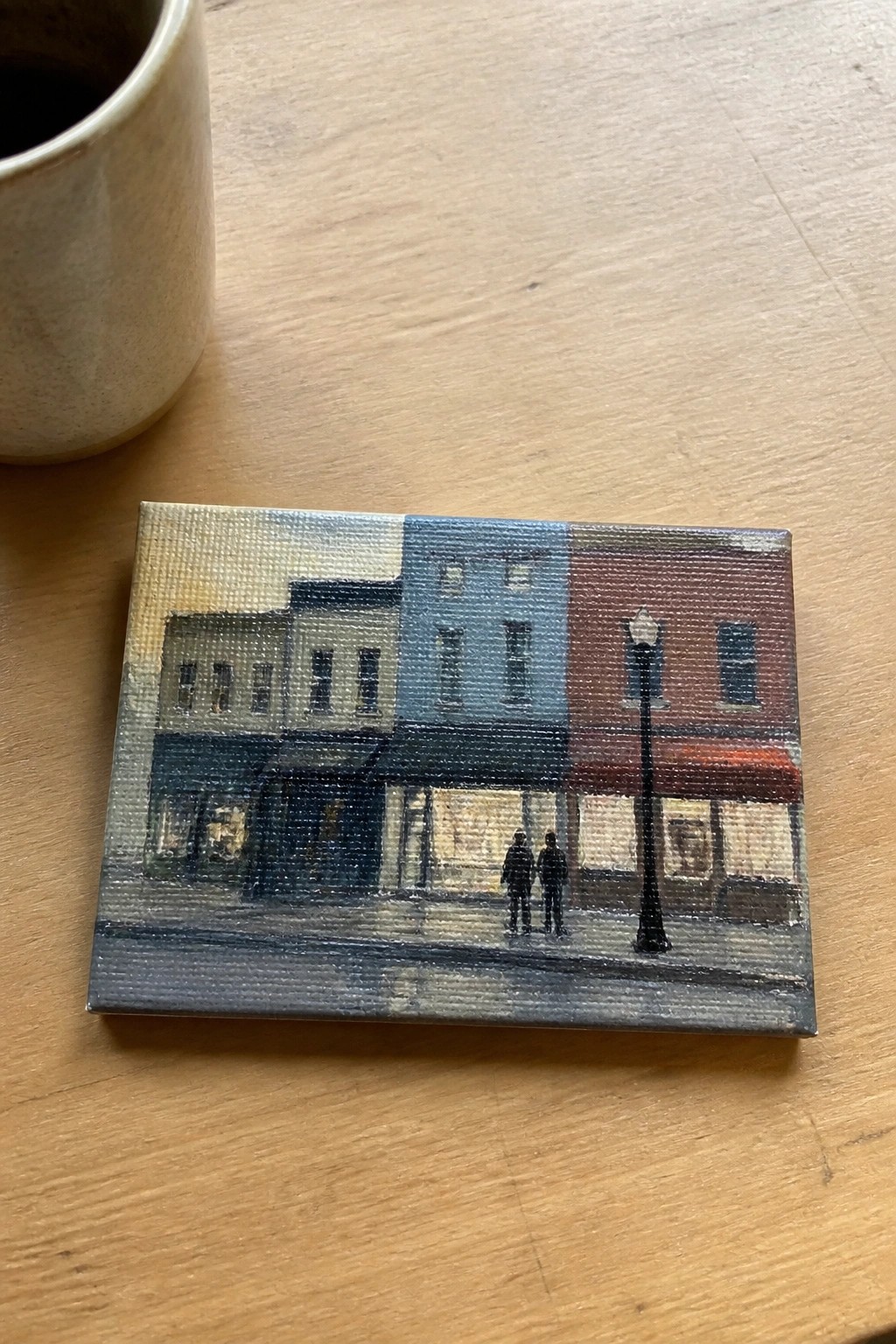 Small canvas painting of colorful buildings, two figures, and lamppost beside beige mug on wood.